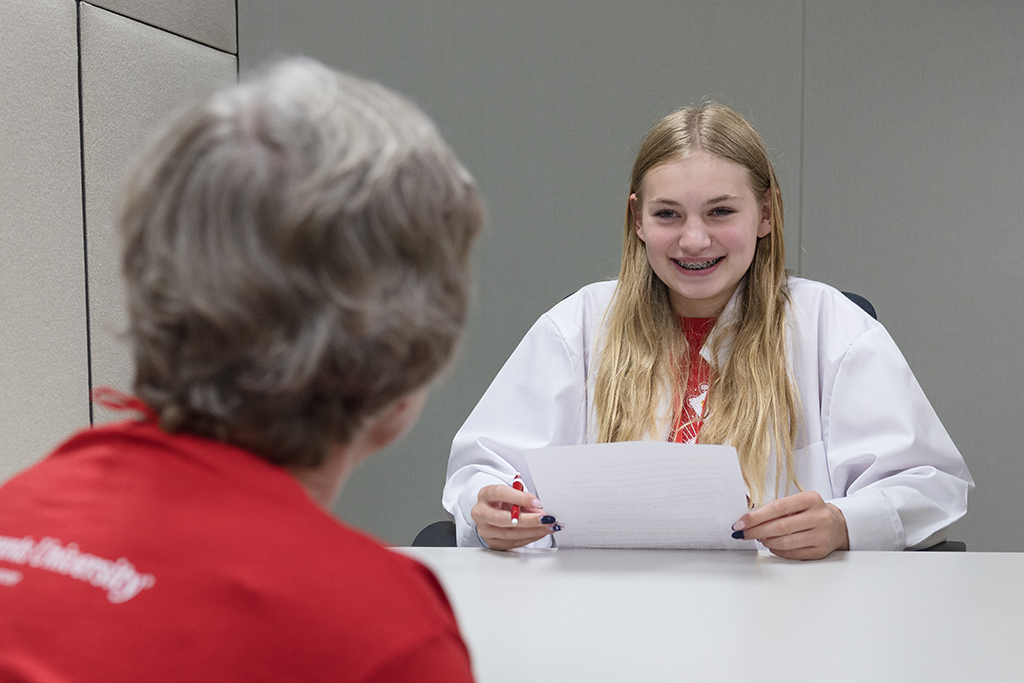 A smiling teenage girl with braces and long blonde hair, wearing a white lab coat, holds a sheet of paper and looks across a table at an adult, who is only visible from the back.