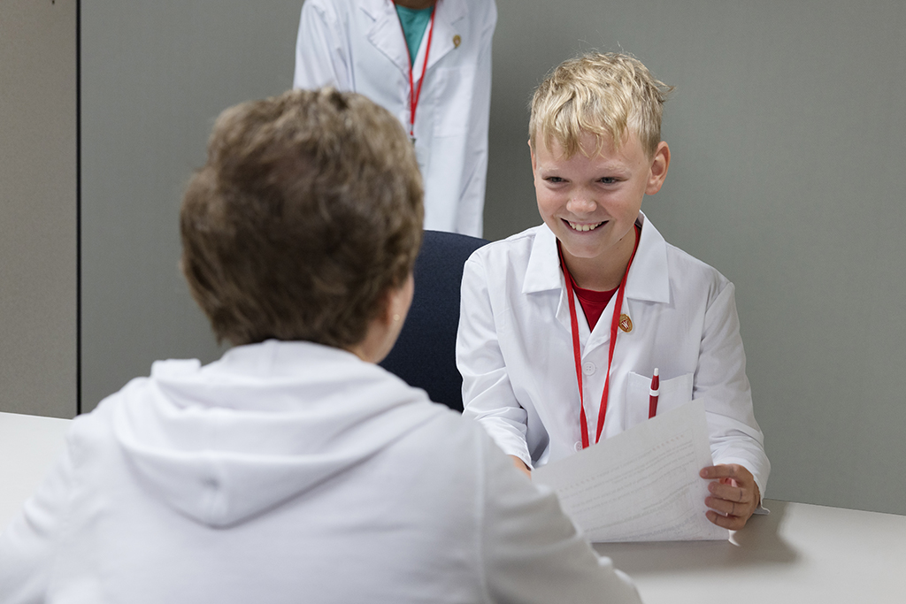 A young boy in a white lab coat with a red lanyard smiles brightly while talking to an adult, who is seated with their back to the camera.