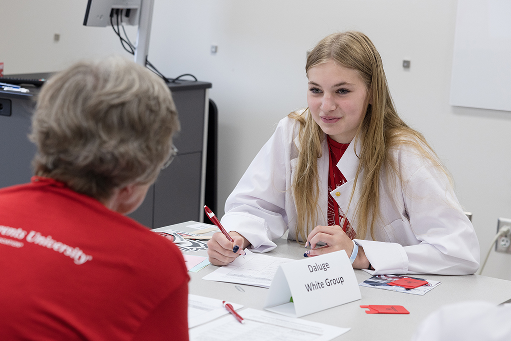 A smiling teenage girl in a white lab coat holds a pen and looks at an older person in a red t-shirt, who is only visible from the back. A name card on the table reads "Daluge White Group."