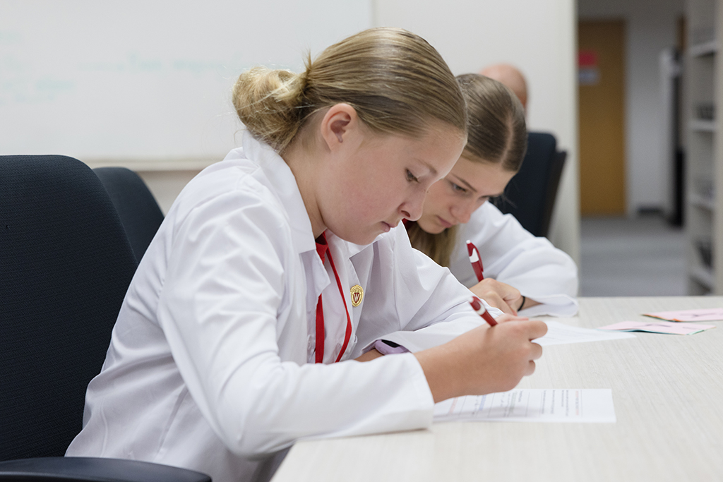 Two girls, one in the foreground, both wearing white lab coats, sit side-by-side at a light-colored table, focused on writing on sheets of paper.