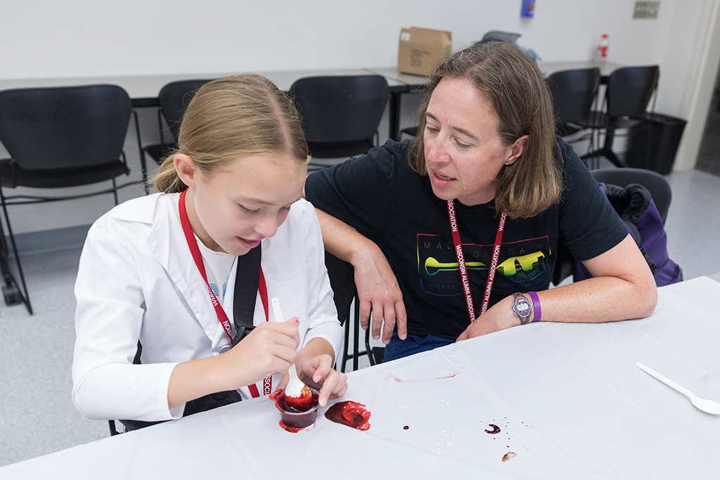 A young girl mixes a red liquid in a cup next to Kim Lintner