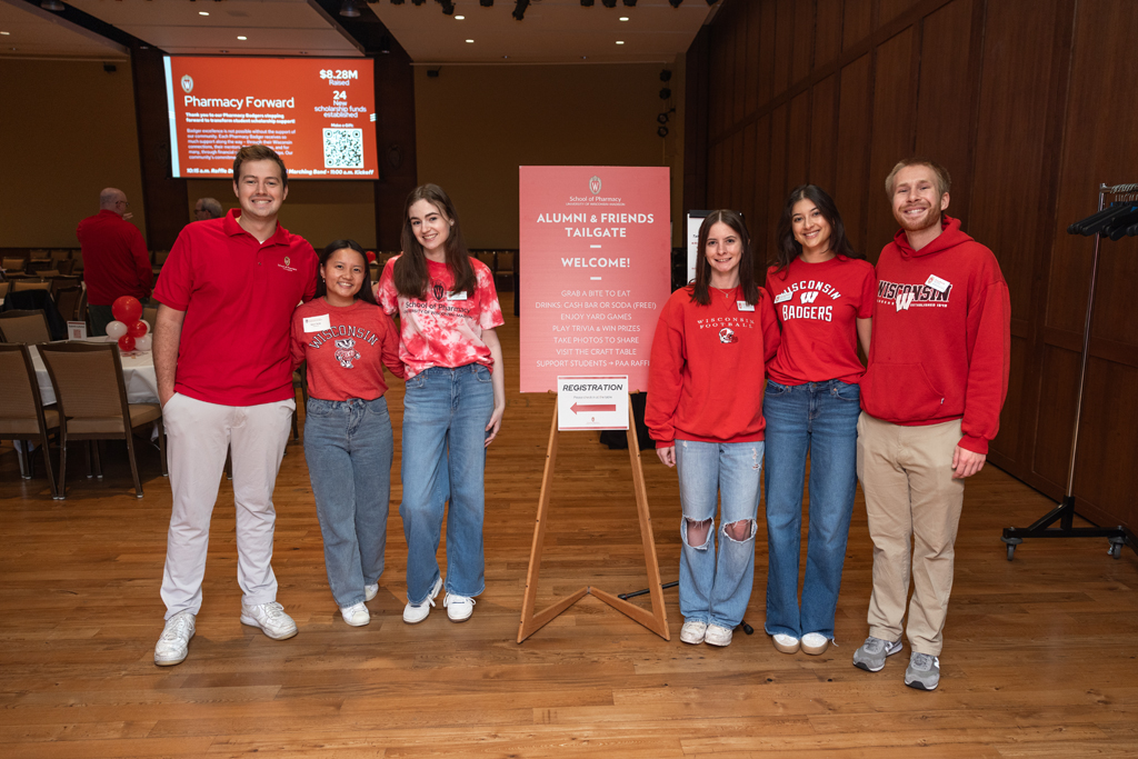 A group of six young adults, all wearing red and white Wisconsin apparel, stand smiling for a photo next to a welcome sign that reads "Alumni & Friends Tailgate."