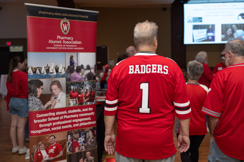 An older man wearing a red Wisconsin Badgers jersey with the number '1' on the back stands facing away from the camera in a crowded event hall, next to a large banner for the Pharmacy Alumni Association.