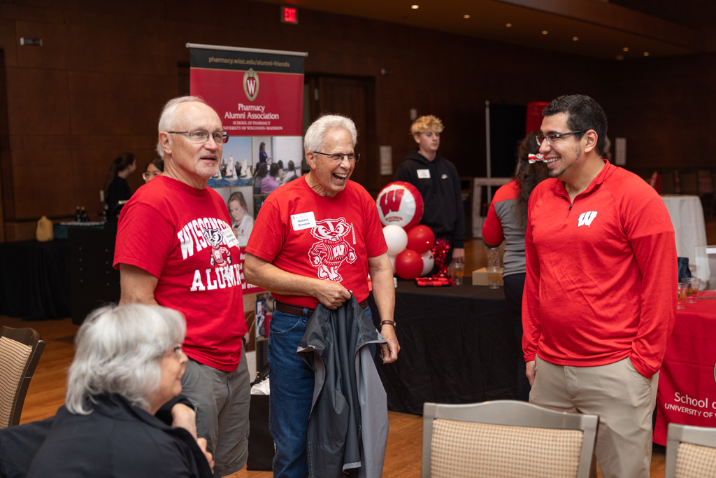 Three men in red Wisconsin shirts are standing and talking animatedly in an event hall, while a woman with gray hair is seated in the foreground.
