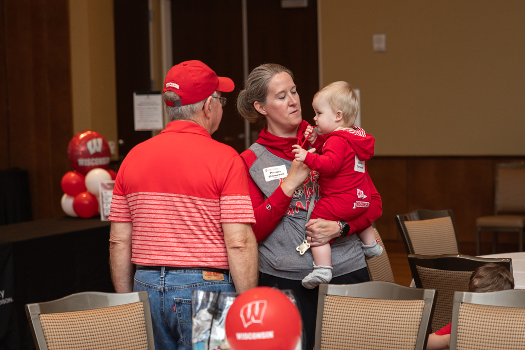 A woman holds a baby in a red outfit while speaking to an older man in a red striped polo shirt and red cap. Red and white balloons are visible in the foreground and background.