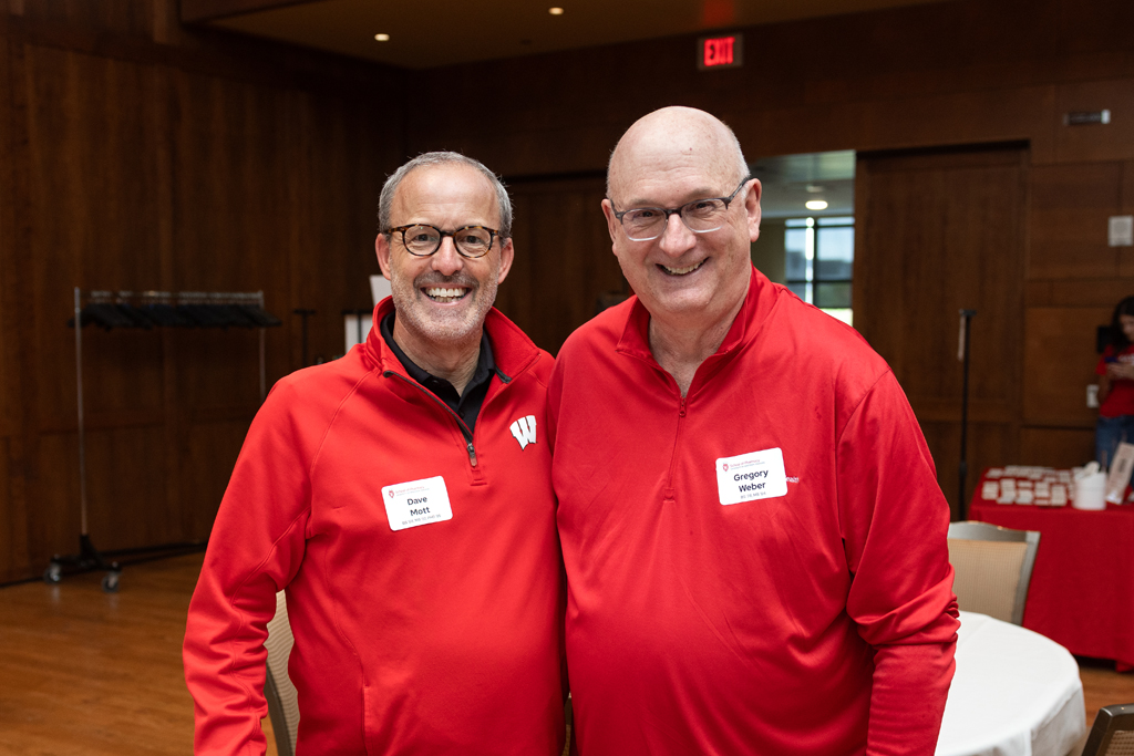 Two men, Dave Mott and Gregory Weber, wearing red zip-up shirts and glasses, are smiling for a photo in an event hall with wood-paneled walls.