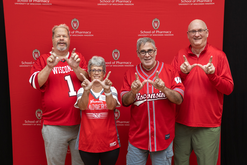 A group of four adults (three men and one woman) are posing in front of a red backdrop with the School of Pharmacy logo. They are all wearing red Wisconsin apparel and making the 'W' hand sign.