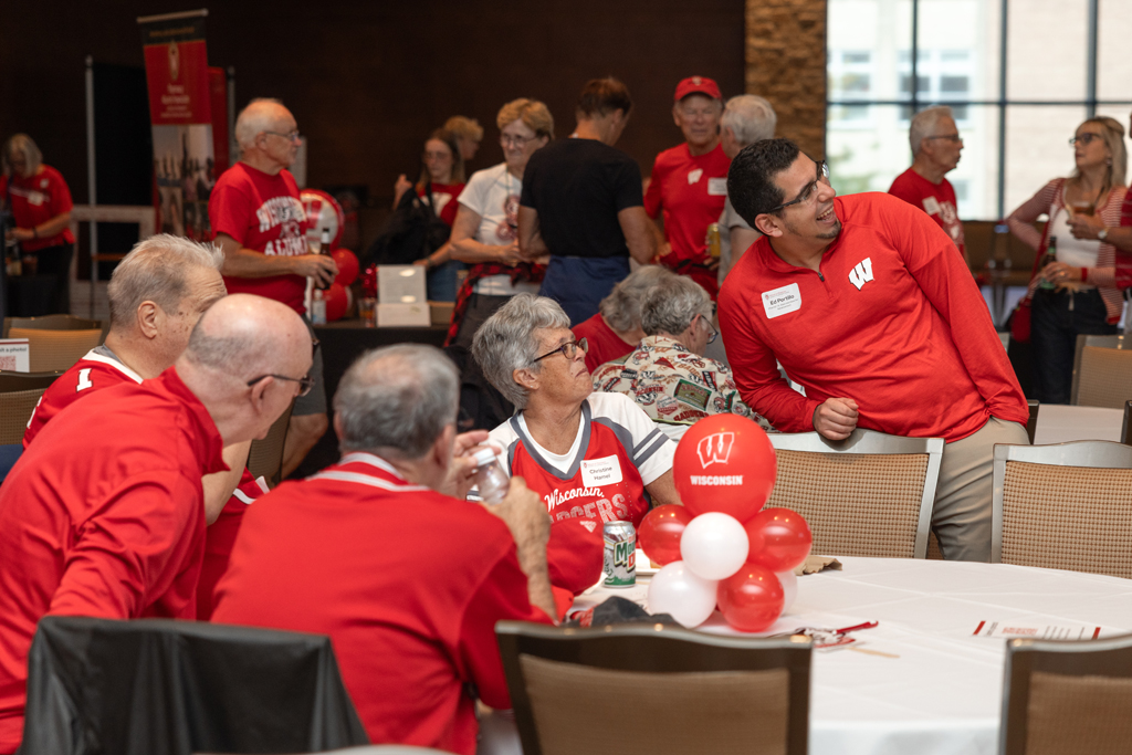 A man in a red shirt is leaning toward a round table, speaking to a group of seated adults, many of whom are wearing red Wisconsin apparel. Red and white balloons are on the table.