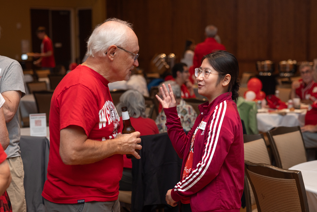 An older man holding a bottle of beer is speaking with a young woman in a red and white striped jacket, who is gesturing with her hand.