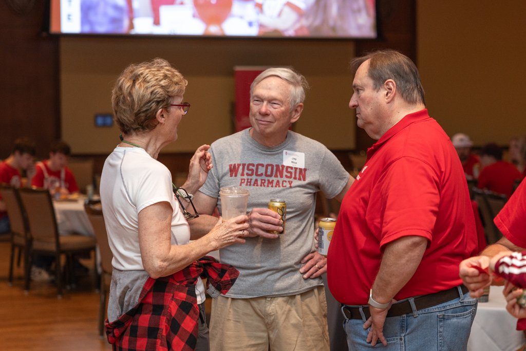 A woman and two men are standing in a small circle, engaged in conversation. The man in the middle wears a gray shirt that says "Wisconsin Pharmacy."