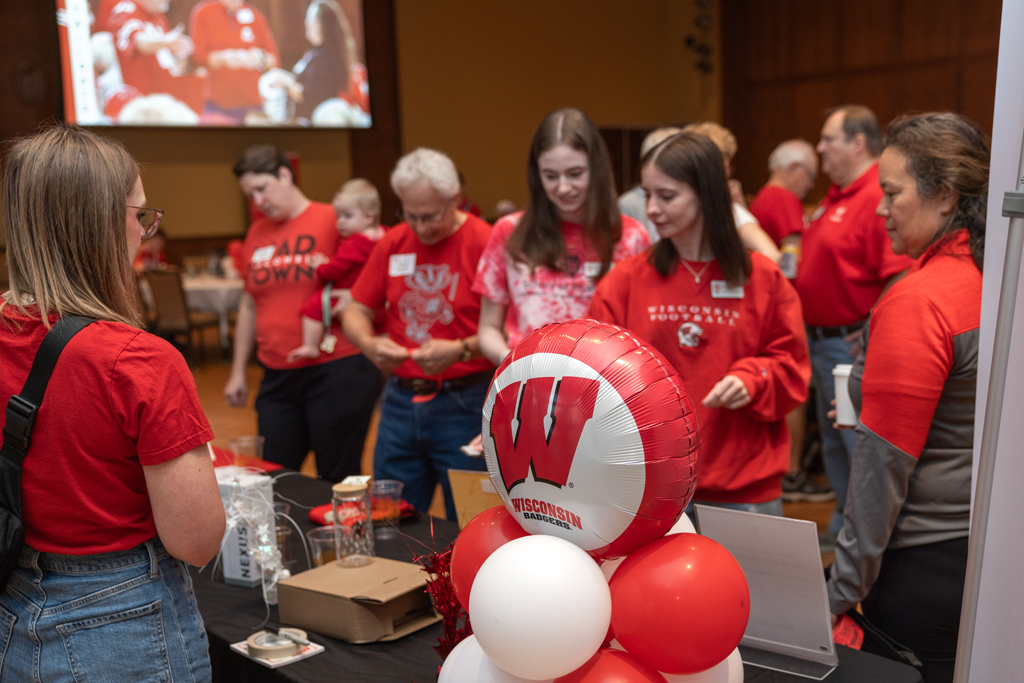 Several people are standing and talking around a table adorned with a large red and white Wisconsin football balloon and other red and white balloons.