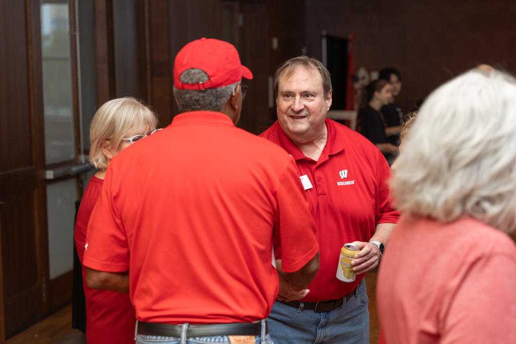 A man in a red polo shirt is smiling and speaking to another man whose back is to the camera in a warmly lit event hall. Both men are wearing red Wisconsin-themed apparel.