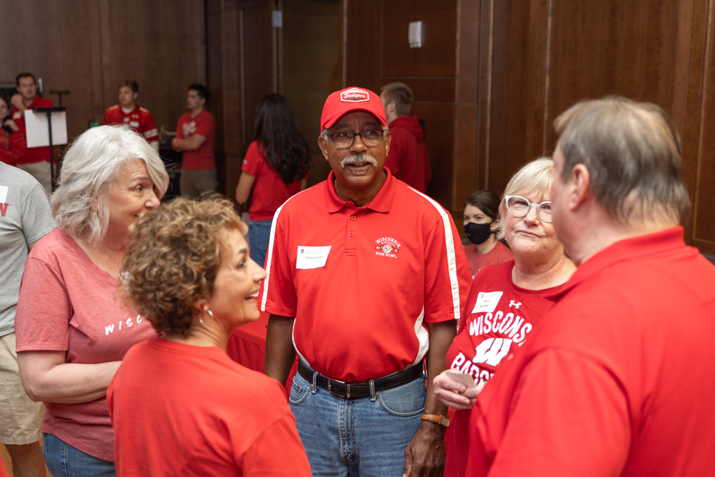 A man in a red polo shirt and cap stands in the middle of a group of four other adults, who are all wearing red or white and engaged in conversation.