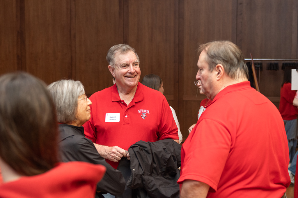 A man in a red polo shirt with a name tag is smiling and speaking to a woman on his left and another man on his right, inside an event room with wood-paneled walls.