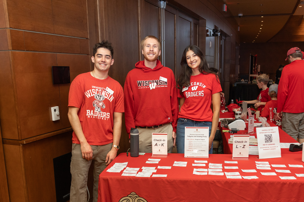 Three young adults, all wearing red Wisconsin shirts, stand smiling behind a check-in table covered with a red cloth. The table has signs for alphabetical check-in sections and numerous name tags.