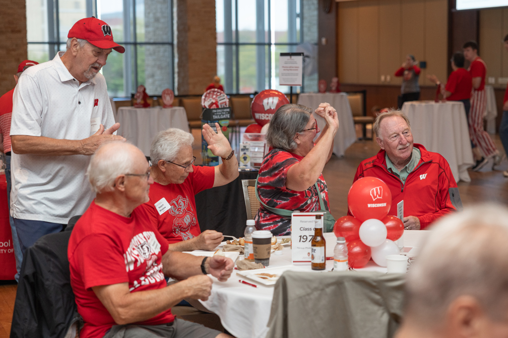 A group of older adults is seated around a table decorated with red and white balloons, and a sign indicating "Class of 1974." Two people are raising their hands as an older man stands nearby.