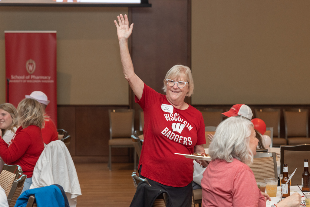 A woman in a red "Wisconsin Badgers" shirt smiles and raises her right hand while standing at a table. She is holding a plate of food.