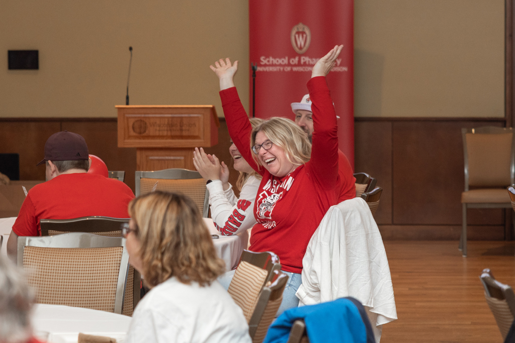 A woman in a red long-sleeve shirt is seated at a table in an event hall, laughing exuberantly with both hands raised in the air.