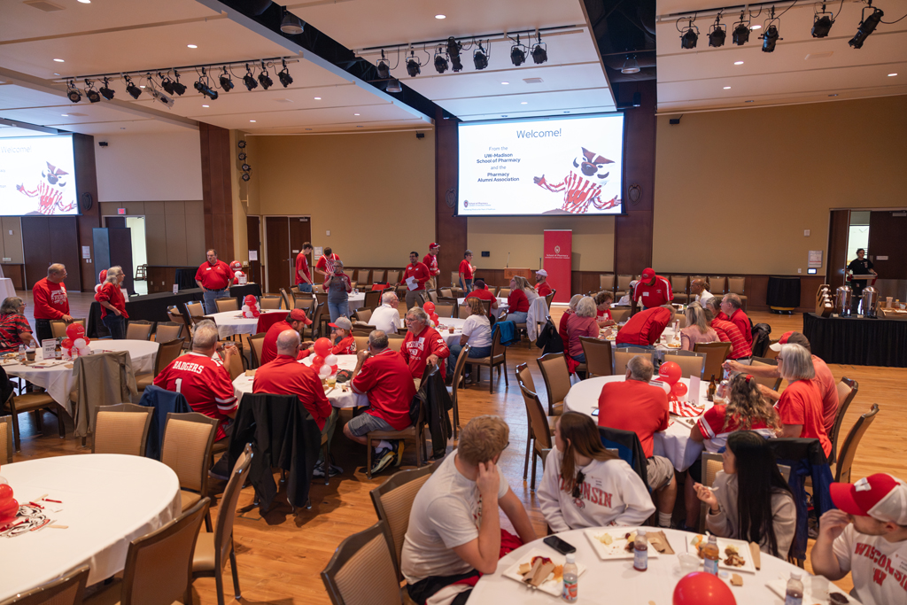 A wide shot of a large, crowded event hall where attendees are seated at round tables. A large screen at the front of the room displays a welcome message and a graphic of Bucky Badger.