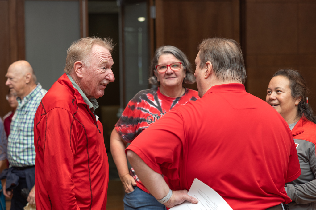 A man in a red jacket is smiling and speaking to a man in a red shirt. Two women are partially visible behind them.