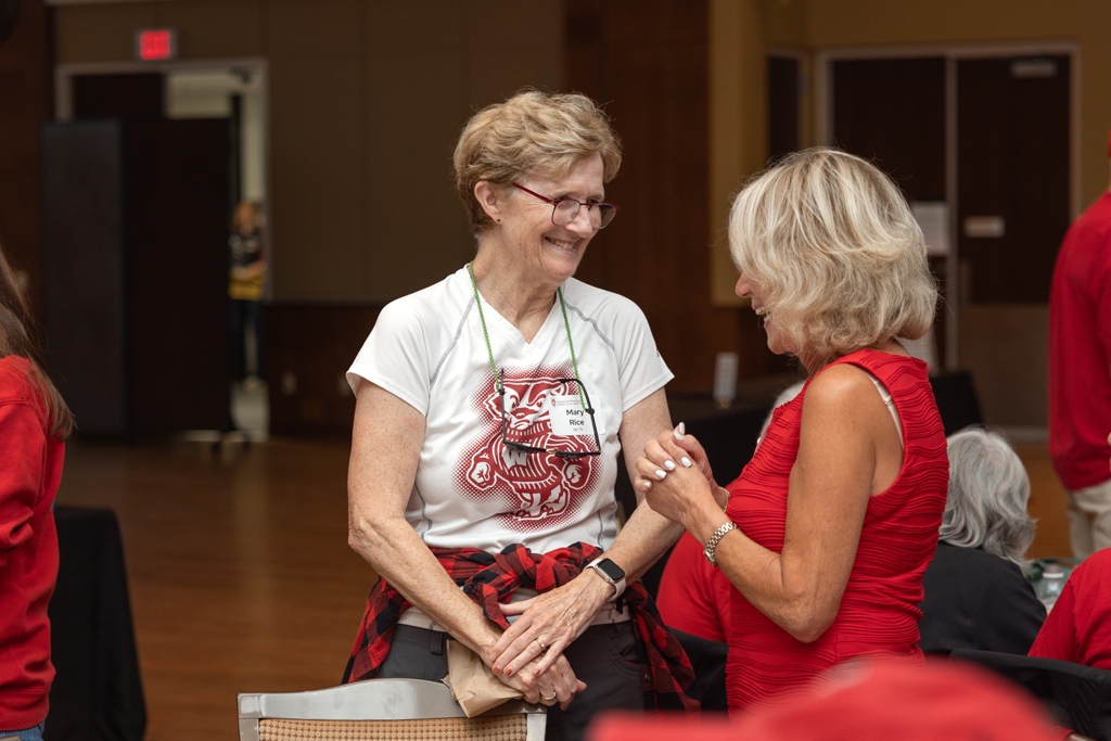 Two women stand facing each other, engaged in conversation. One wears a white Bucky Badger shirt with a plaid shirt tied at her waist, and the other wears a red tank top.
