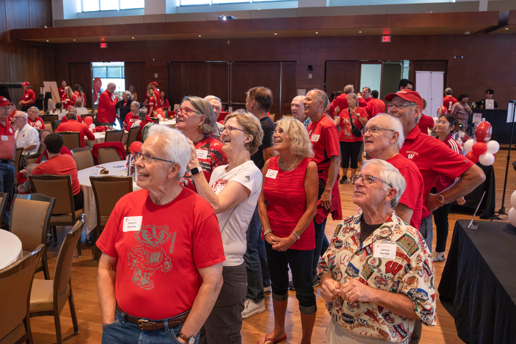 A large group of adults, most wearing red University of Wisconsin apparel, stand in an event hall, looking up and smiling.