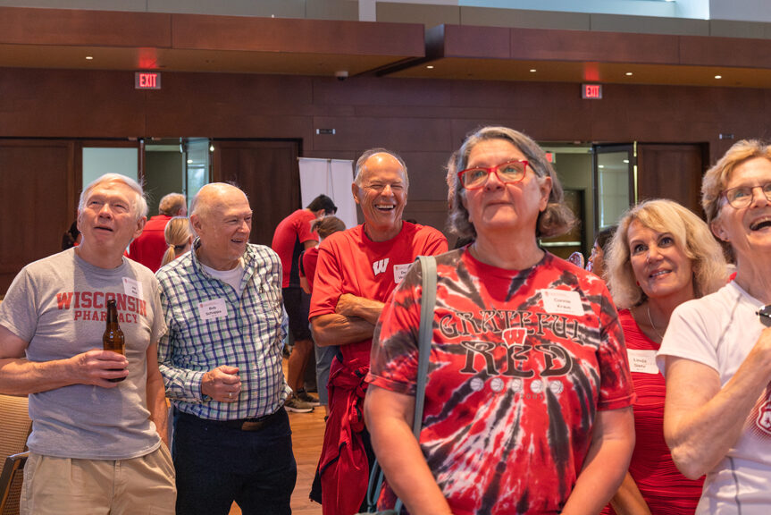 A close-up of five adults standing together in an event hall, smiling and laughing. They are wearing a mix of red, white, and tie-dye Wisconsin-themed clothing.