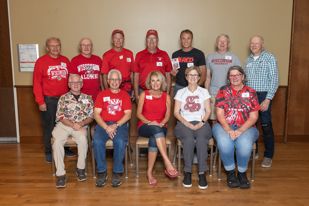 A group portrait of twelve adults (ten men and two women) posed in two rows. Most are wearing red Wisconsin-themed apparel, standing and sitting against a neutral tan wall.
