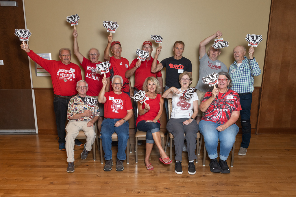 A group portrait of twelve adults holding up small Bucky Badger signs. They are smiling and posed in two rows against a tan wall.