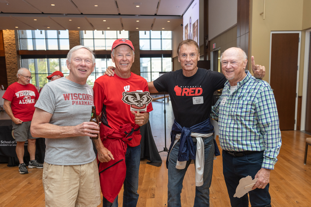 Four men are standing together in a brightly lit event hall, smiling for a photo. They are wearing red, gray, and plaid shirts.