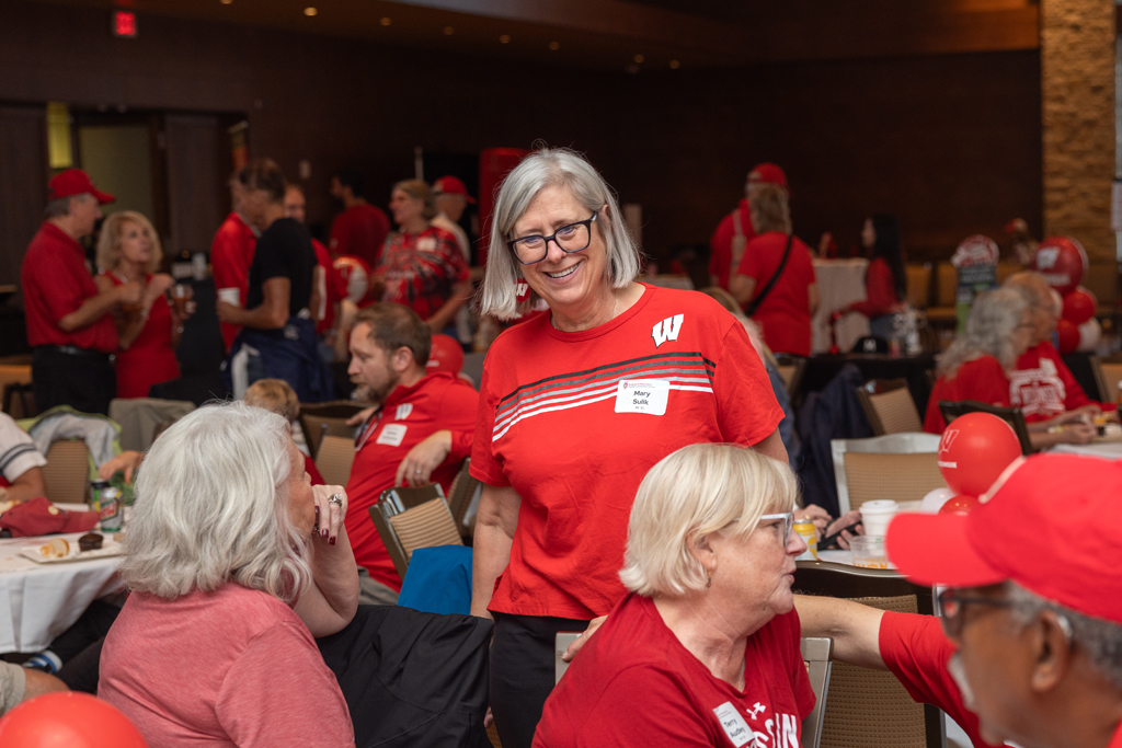 A woman in a red shirt with a white 'W' logo stands in the center of a crowded event room, smiling at the camera. Other attendees are seated at tables around her.