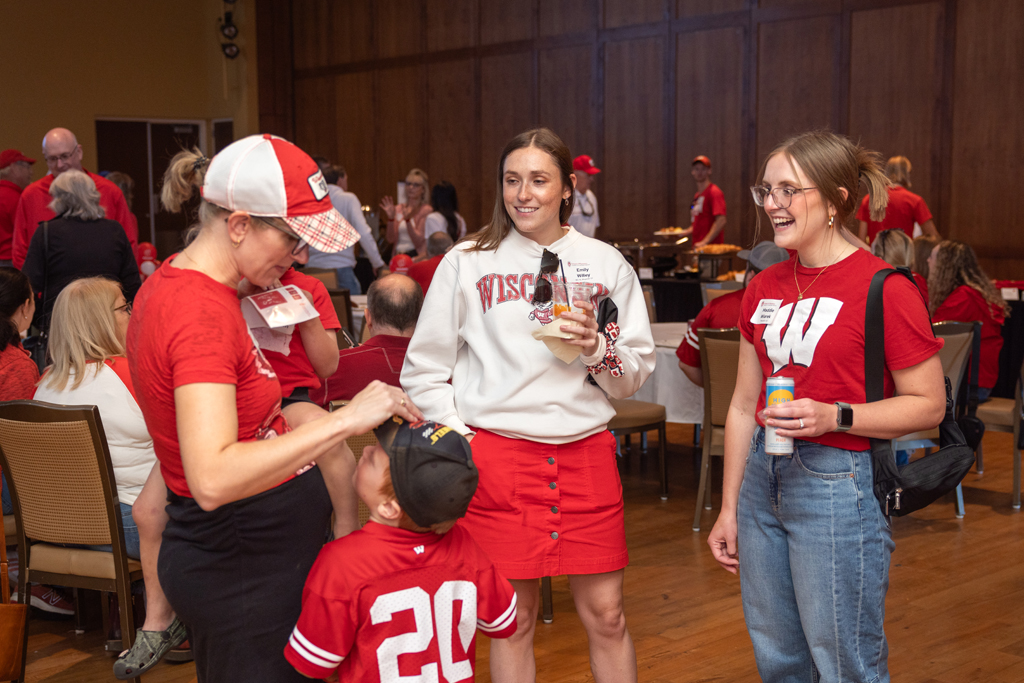 Two women and a young boy in a red jersey are standing and conversing in an event hall. The woman on the right is laughing, and they are all wearing red and white Wisconsin apparel.