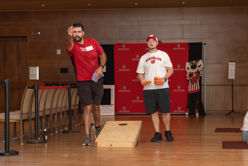 Two men are playing Cornhole (beanbag toss) on a wooden floor indoors. One man is aiming a toss while the other holds beanbags. A red School of Pharmacy backdrop is visible behind them.