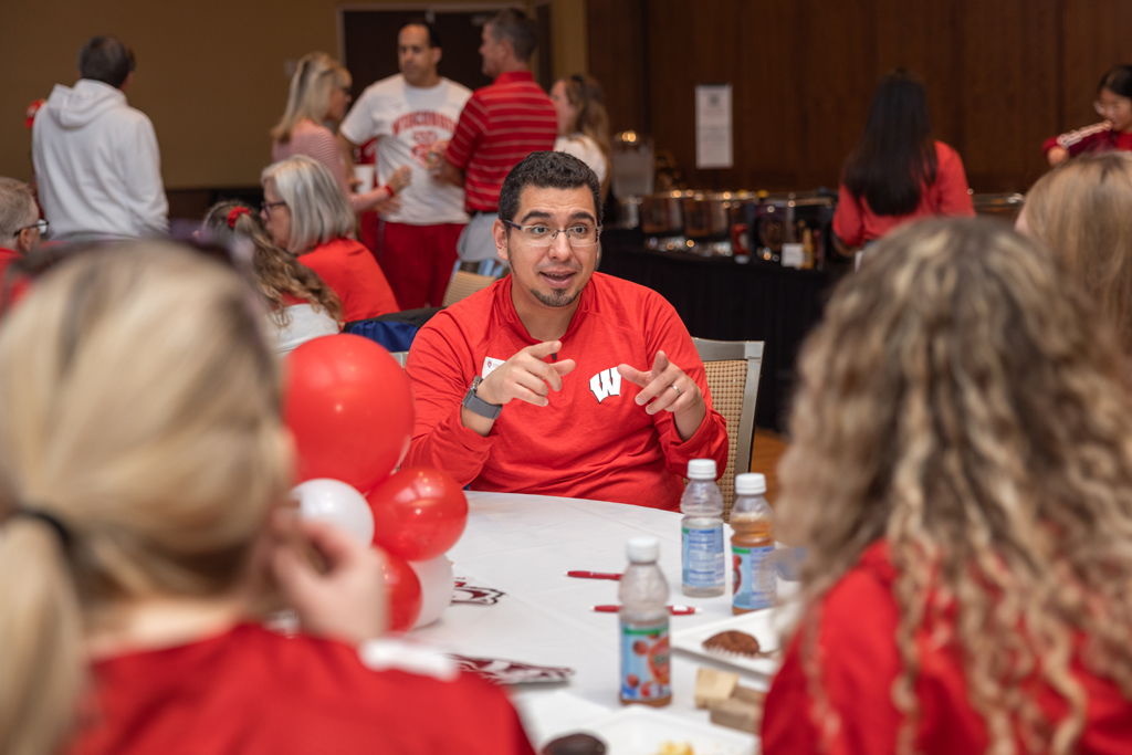 A man in a red Wisconsin sweatshirt is seated at a table with a white cloth, gesturing with his hands while speaking to unseen people around the table. Red and white balloons and drinks are visible.