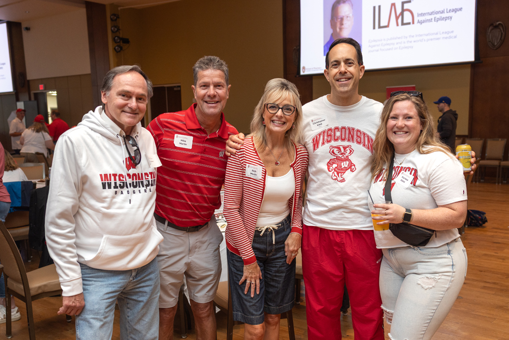 Five adults stand together and smile for a photo in an event hall. They are wearing a mix of red and white Wisconsin apparel. A projection screen with the ILAE (International League Against Epilepsy) logo is in the background.