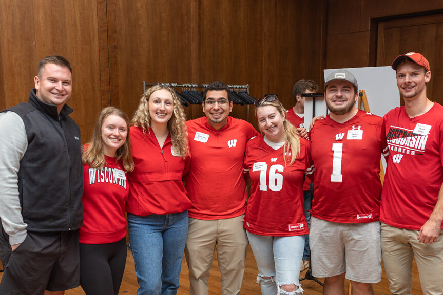 A group of seven young adults, most wearing red University of Wisconsin shirts or jerseys, are standing together and smiling at the camera in a wood-paneled event room.
