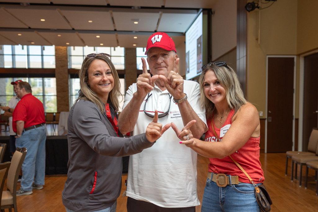 Two women and an older man in a red cap are standing together, smiling, with all three people making the 'W' hand sign. The background is a large indoor event space.
