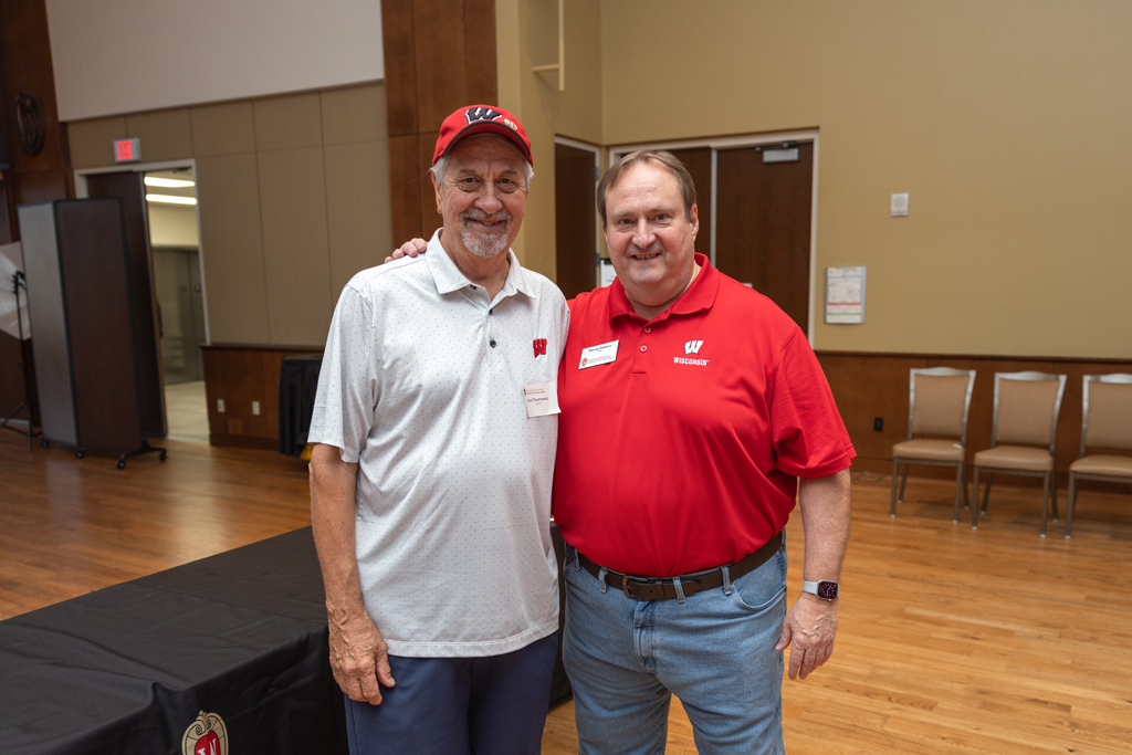 Two men stand together in an event hall with light brown walls and wooden floors. The man on the left wears a white polo and a red cap, and the man on the right wears a red polo shirt. They are both smiling.