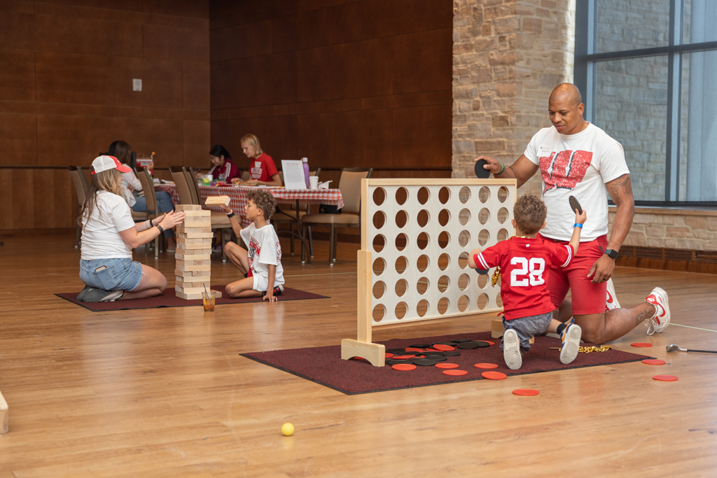 A group of adults and children are playing oversized indoor games on a wooden floor. In the foreground, a man and a young boy play Connect Four, while a woman and another boy play Jenga in the middle ground.