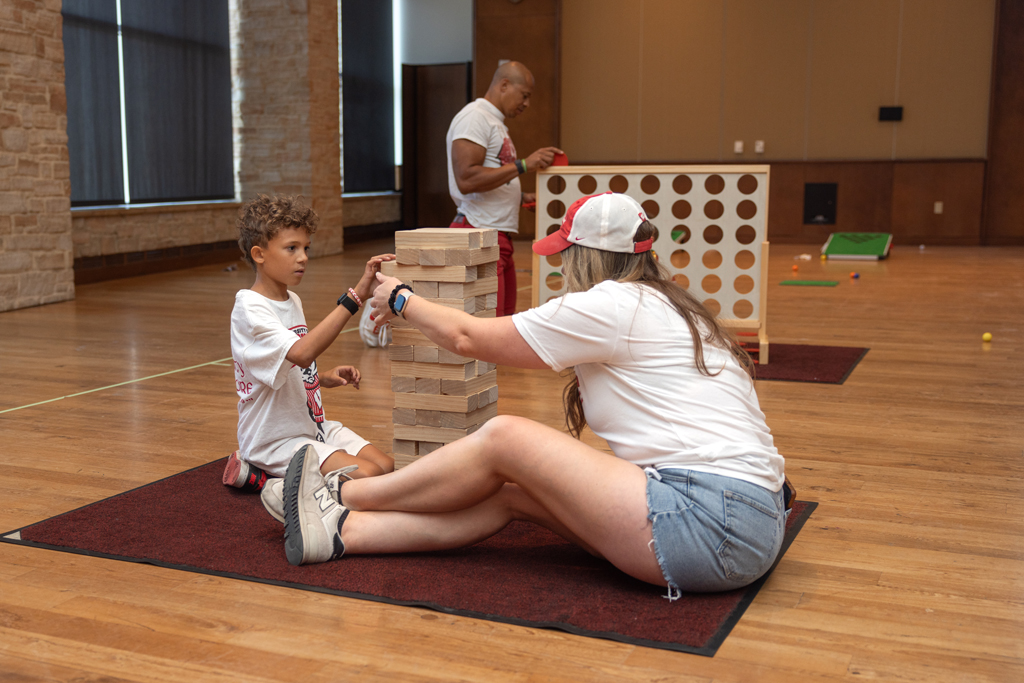 A young boy and a woman are seated on the floor, playing a game of large-block Jenga indoors. A man is standing in the background near a large Connect Four game.