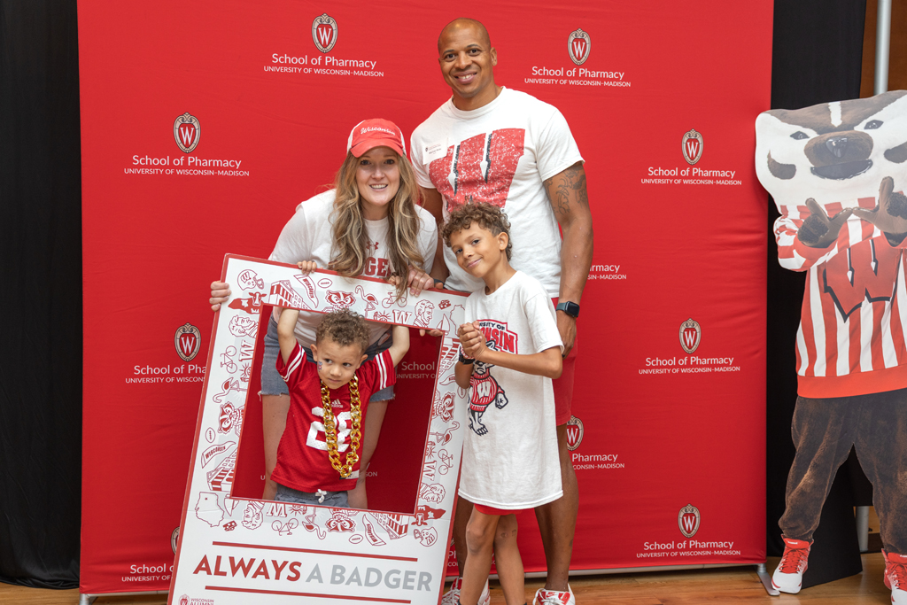 A family of four is posing in front of the red School of Pharmacy backdrop. One young boy is framed in a large photo prop that says "ALWAYS A BADGER." A Bucky Badger cardboard cutout is partially visible on the right.