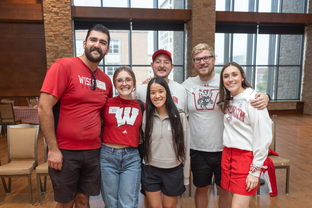 A group of six young adults stands together and smiles for a photo in a large, windowed event space. They are all wearing casual red, white, and gray University of Wisconsin apparel.
