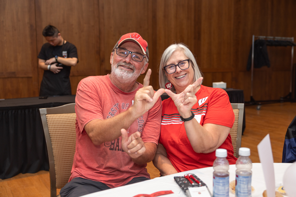 A man wearing a red cap and shirt, and a woman wearing a red and white Wisconsin shirt, are seated at a table and smiling. Both are making the 'W' hand sign.