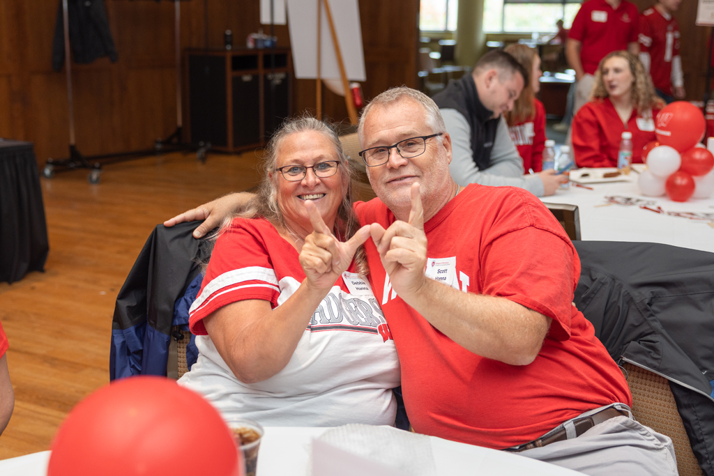 A man in a red shirt and a woman in a white and red shirt are seated at a table, smiling at the camera. Both are making the 'W' hand sign.