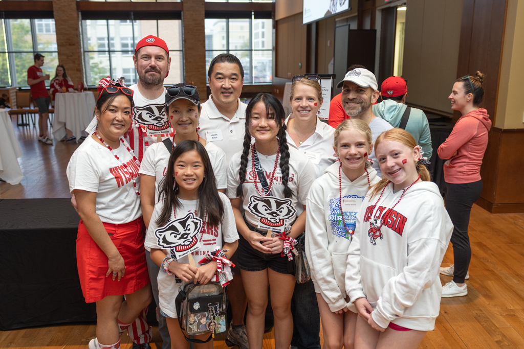 A large group of nine people, including adults and children, are posing together and smiling for a photo in an event space with large windows overlooking a city street. Most are wearing red and white Wisconsin-Badger themed clothing.