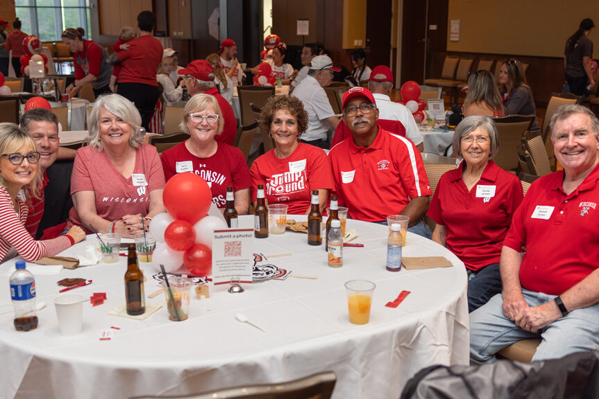 Seven adults, most wearing red and white Wisconsin attire, are seated around a round table with a white tablecloth in a large event room. They are smiling at the camera, with red and white balloons and drinks on the table.