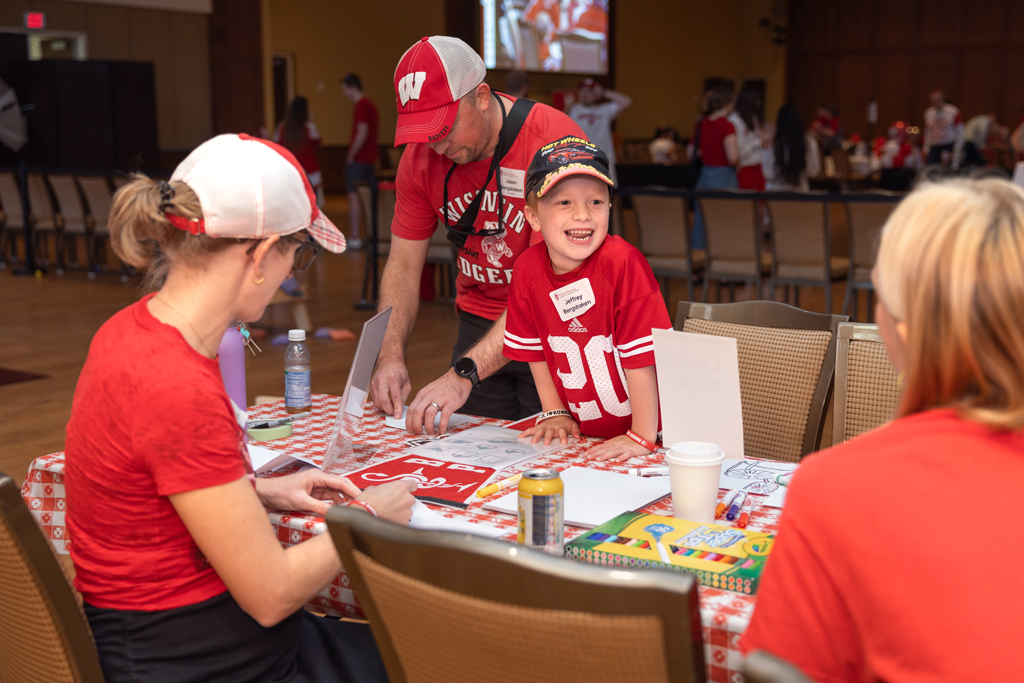 A young boy in a red jersey (number 20) smiles widely as he stands at a table with a red and white checkered tablecloth. A man in a red cap is standing next to him, and a woman is seated in the foreground.