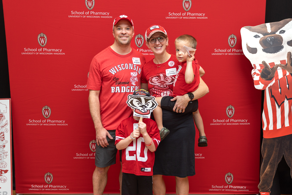A family of four, including two adults and two young boys wearing red University of Wisconsin-Madison apparel, poses in front of the red School of Pharmacy backdrop. One child is held by a parent, and the other holds a Bucky Badger sign over his face.