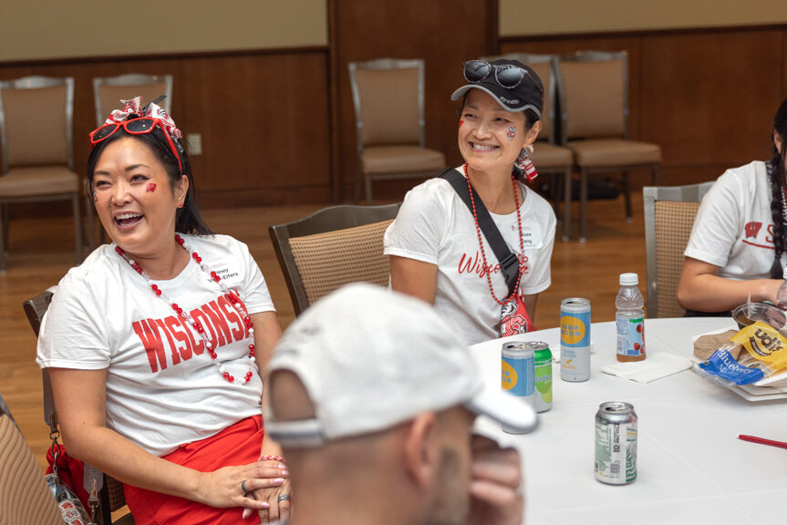A candid image of two women and a girl seated at a table at an indoor event. They are all wearing red and white Wisconsin gear and smiling or laughing.
