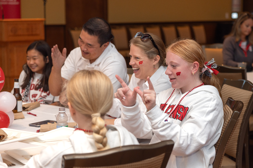 A candid shot of a group of people seated around a table, with a red-haired girl in the foreground making the 'W' hand sign. Other people at the table in the background are smiling and talking.